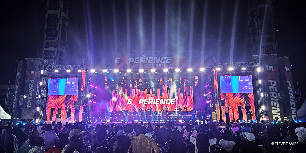 Wide view of The Experience 2025 in Lagos, Nigeria, showing a large outdoor stage with lighting and video screens performing before a crowd of worshippers during the annual gospel music event.
